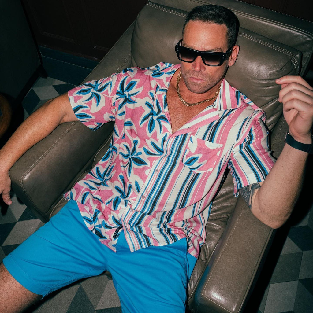 man in designer sunglasses sitting on a couch wearing a blue and pink tropical short sleeve button down shirt and blue shorts